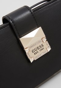 Musta nahkalaukku, jossa on näkyvä kiiltävä kultainen solki, jossa on niittejä ja kristalleja, kaiverrettu logo, jossa lukee "Guess EST. 1981."