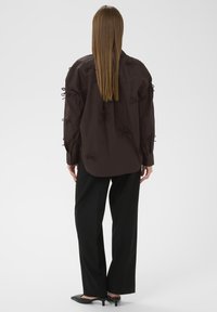 Mørkebrun bluse med dekorative sløjfe-detaljer på ærmerne, parret med sorte brede bukser og spidse sorte hæle. Minimalistisk design.