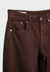 Tmavě hnědé džíny z denimu s předními kapsami, poutky na pásek, stříbrnými nýty a zapínáním na knoflík, zobrazující pohled zblízka na horní oblast pasu.