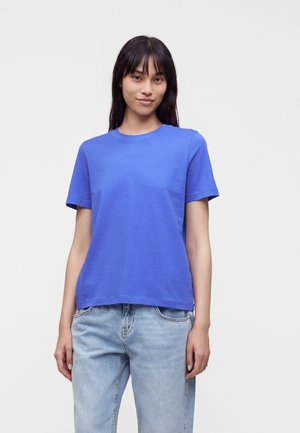 Jeune femme portant un t-shirt bleu uni à manches courtes et un jean bleu clair, debout devant un fond clair et uni.