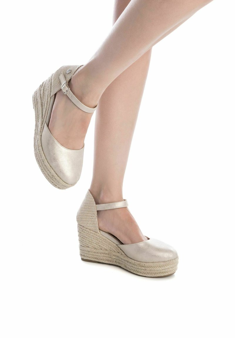 Sandalias de cuña beige con un acabado metálico, plataforma de yute tejido y correa al tobillo. Superficie suave con detalle texturizado en el tacón.