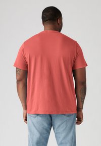 Kortærmet, koralfarvet T-shirt, lavet af bomuldsstof, afslappet pasform, rund halsudskæring, glat tekstur, båret med lyseblå jeans, synlig tatoveringsdetalje.