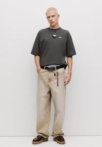 Grå oversized t-shirt, løse beige jeans med et dekorativt bælte, robuste brune sko og lagede sølvhalskæder. Enkle teksturer og farver.