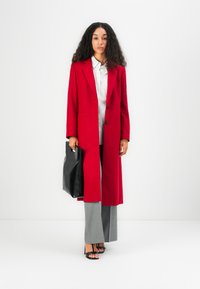 Manteau en laine rouge à coupe droite, fermeture à un bouton, associé à un pantalon large gris et un sac à main en cuir noir.
