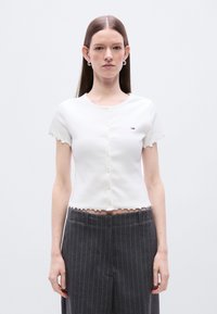 Λευκό cropped cardigan με κυματιστές άκρες και κοντά μανίκια, με κουμπιά μπροστά και μικρό λογότυπο στο στήθος.