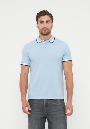 Άνδρας που φορά ανοιχτό μπλε πόλο με σκούρες λεπτομέρειες στον γιακά και τα μανίκια, συνδυασμένο με σκούρο γκρι τζιν, στέκεται μπροστά σε λευκό φόντο.