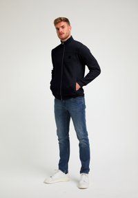 Tamsiai mėlyna zip-up striukė su struktūruota medžiaga, aukštu apykakle ir šoninėmis kišenėmis, derinama su mėlynomis džinsais ir baltais sportiniais bateliais.