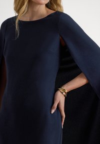 Nāvijas zilas krāsas kleita ar apmetņa stila piedurknēm, ar gludu tekstūru un smalku sānu kabatas detaļu. Uz plaukstas ir zelta aproču rokassprādzes.