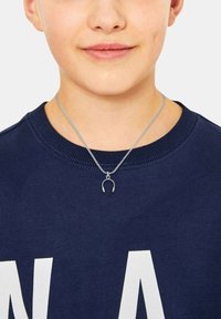 Zilveren metalen ketting met een kleine, gedetailleerde hanger in de vorm van een hoofdtelefoon, gedragen over een donkerblauwe sweater met witte tekst.