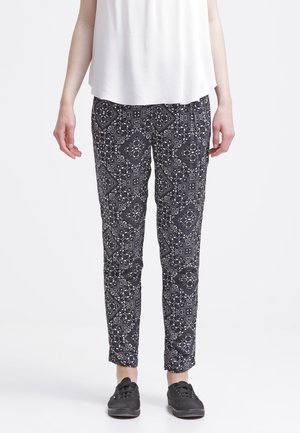 Personne portant un pantalon slim à motif noir et blanc, un haut blanc sans manches et des chaussures noires à lacets, debout sur un fond blanc.