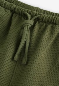 Olivovo zelené šortky vyrobené z textúrovanej tkaniny s pásom na šnúrku a uzlovým detailom. Majú mäkkú, priedušnú textúru.
