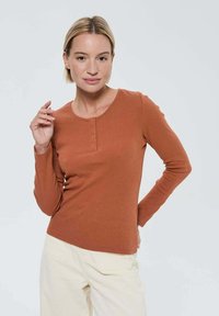 Femme aux cheveux blonds courts portant une chemise à manches longues couleur rouille avec des boutons et un pantalon beige clair, posant avec une main près de son visage.