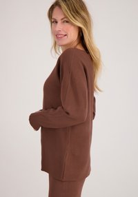 Bruine trui van zacht materiaal, met een ronde hals, een ontspannen pasvorm en geribbelde manchetten. De zoom is iets langer aan de achterkant.