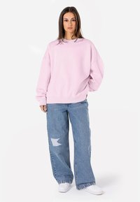 Sweatshirt oversize rosa chiaro con polsini a costine, abbinato a jeans a gamba larga azzurri chiari con un ginocchio strappato. Le sneakers bianche completano il look.