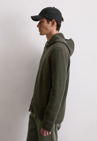 Hooded zip-up jacket in olive green fabric, featuring ribbed texture, a relaxed fit, and a visible zipper. Black cap adds contrast.
