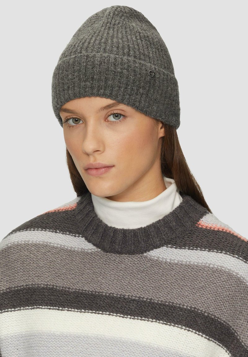 Grijze geribbelde breimuts met een licht puntige top, voorzien van een klein logoaccent. Model draagt een gestreepte trui eronder.