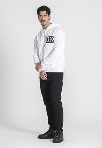 Sudadera blanca con logo negro "GK", combinada con jeans negros y zapatillas negras. El modelo está de pie con los brazos relajados, contra un fondo neutro.