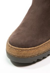 Brązowy zamszowy botek do kostki z teksturowaną cholewką, kontrastowym brązowym wewnętrznym wykończeniem z wełny oraz grubą czarną gumową podeszwą, z widocznymi detalami szycia.