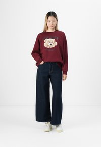 Woman standing with hands in pockets wearing burgundy sweatshirt with teddy bear graphic and dark wide-leg jeans against white background.