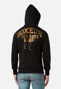 Felpa nera con cappuccio, decorata con una stampa grafica dorata sul retro, che presenta il testo "BOXEUR DES RUES" e una figura in posa da boxe.