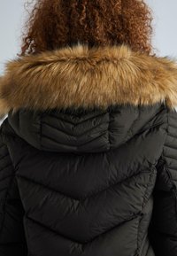 Cilvēks ar lokainiem matiem, ģērbts melnā daudzkārtīgā jakā ar brūnu mākslīgā apdaringa kapuci, skatīts no aizmugures.