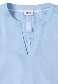 Blouse bleu clair à col en V, détail froncé, avec des étiquettes de marque indiquant "CECIL" et "seaside". Texture de tissu lisse.