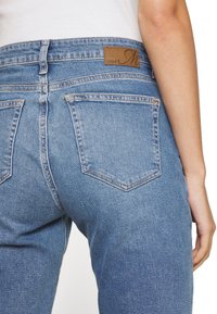 Jeans de mezclilla de color azul claro con cintura alta, que cuentan con una parche de cuero marrón, bolsillos traseros con un diseño cosido y un ligero desgaste.