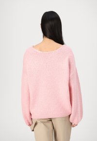 Pullover lavorato a maglia oversized di colore rosa con scollatura ampia e maniche a palloncino, caratterizzato da una consistenza morbida e orlo a coste. Indossato con pantaloni beige.