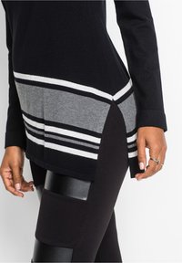 Černý svetr se šedobílým proužkovaným panelem a bočními rozparky, nošený přes černé legíny s lesklými černými záplatami na stehnech.