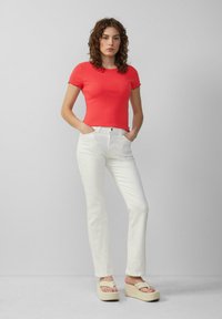 Vrouw staat met handen in de zakken, gekleed in een felrode, getailleerde top met korte mouwen, witte rechte broeken en beige platform sandalen tegen een grijze muur.