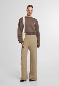 Felpa marrone corta con orlo a coste, abbinata a pantaloni a gamba larga beige in maglia. Il modello tiene una borsa beige a tracolla. Occhiali rettangolari trasparenti.