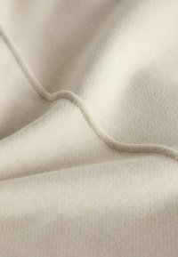 Hellbeige Stoff mit einer glatten, leicht strukturierten Oberfläche, die sanfte Falten und einen subtilen Glanz zeigt und die Nähte betont.