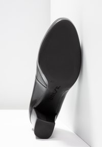 Chaussure à talon haut en cuir noir, inclinée contre un coin blanc, montrant une semelle lisse et un talon carré avec la marque "unisa" et la taille 37.