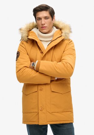 Parka de invierno amarilla con capucha forrada de piel, cierre de cremallera y botón, dos bolsillos de parche, puños acanalados y un pequeño parche de logo en la manga.