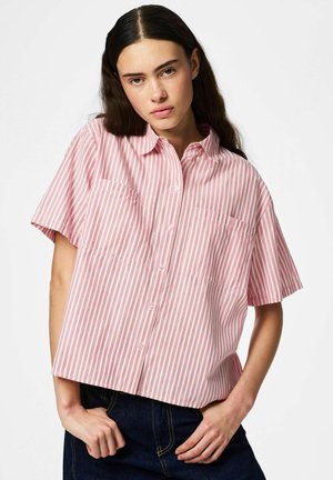 Jonge vrouw die een loszittend roze-wit verticaal gestreept overhemd met korte mouwen en donkerblauwe spijkerbroek draagt, poserend met haar handen vlak bij haar taille.