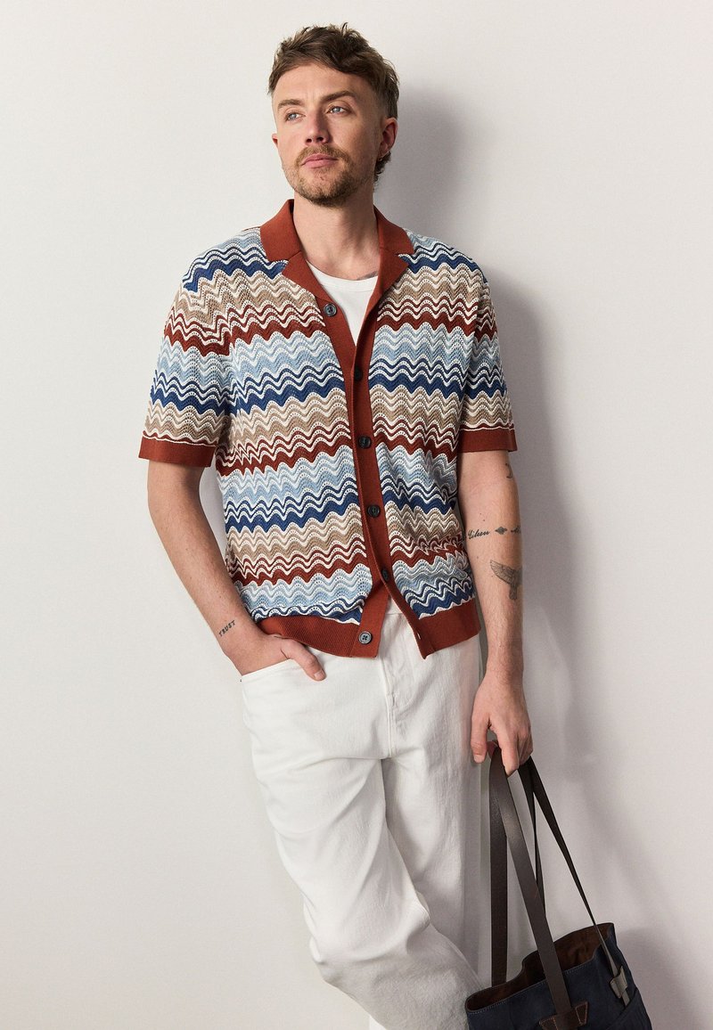 Homme portant un cardigan en maille à manches courtes avec des rayures ondulées bleues, marron et beige, pantalon blanc, tenant un sac cabas bleu marine avec des anses en cuir.
