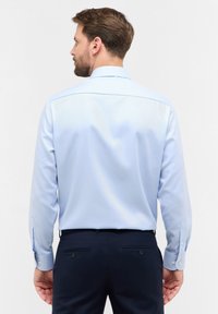 Camicia a maniche lunghe azzurro chiaro con colletto a punta, polsini con bottoni e una texture liscia. Indossata con pantaloni scuri e mostrata da dietro.