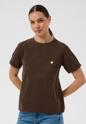 Žena s tmavými vlasmi v cope, oblečená v hnedé tričko s vyhrnutými rukávmi a malým žltým srdiečkom na hrudi, modré rifle, ruky za chrbtom.