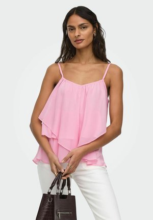 Frau trägt ein mehrlagiges, ärmelloses rosa Oberteil und weiße Hose, hält eine dunkel strukturierte Handtasche mit beiden Händen.