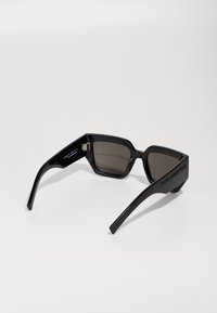 Lunettes de soleil rectangulaires noires avec des verres foncés, dotées d'une épaisse monture en plastique et de branches élégantes et angulaires. Détail de logo notable sur les branches.
