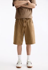 Pantalones cortos de mezclilla marrones con corte recto, con trabillas para el cinturón y bolsillos delanteros. Combinados con una camisa marrón y zapatillas blancas con detalles de color.