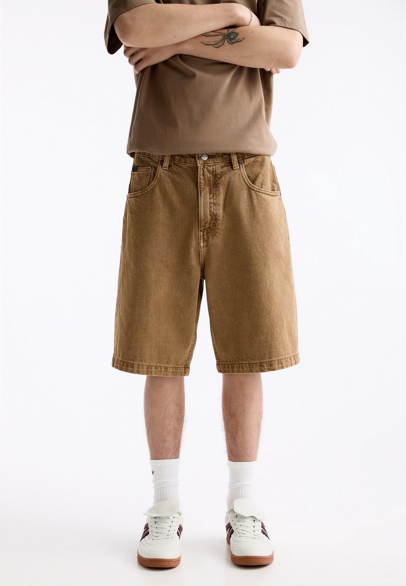 Pantalones cortos de mezclilla marrones con corte recto, con trabillas para el cinturón y bolsillos delanteros. Combinados con una camisa marrón y zapatillas blancas con detalles de color.