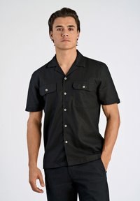 Chemise noire à manches courtes avec un design à col, deux poches avant et boutons métalliques. Texture lisse, coupe ajustée, look minimaliste.