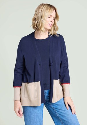 Woman with shoulder-length blonde hair wears navy cardigan with beige pockets and cuffs over dark top and blue jeans, posing with head turned.
