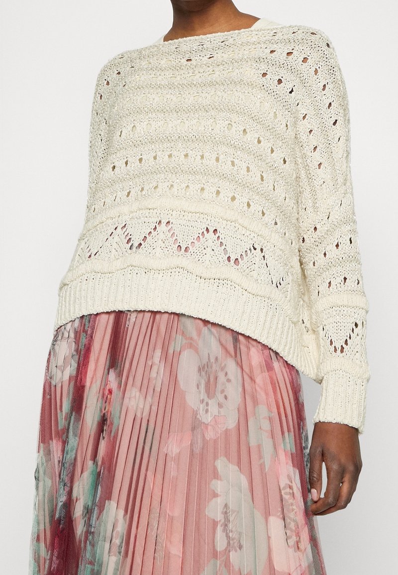 Pull en tricot crème à coupe ample, motif à maille ouverte et ourlet côtelé. Associé à une jupe plissée à fleurs roses aux couleurs douces.