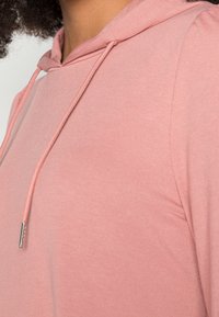 Lichtroze hoodie met een trekkoordkap, een zachte textuur en een getailleerde pasvorm. De stof lijkt soepel en lichtgewicht, geschikt voor het laagjes dragen.