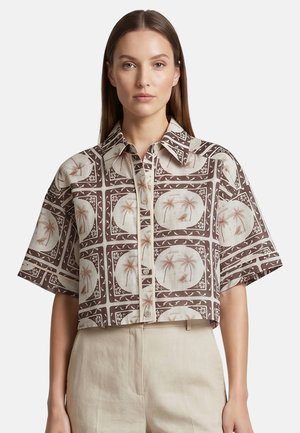 Donna che indossa una camicia cortissima con maniche corte e bottoni, con motivi di palme marroni e beige e forme geometriche.