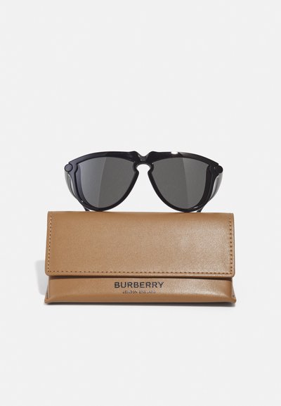 Juodos didelės katės akių formos saulės akinius, padėtus ant rusvo odinio dėklo. Dėkle iškilmingai išspaustas "BURBERRY" logotipas. Lygi tekstūra.