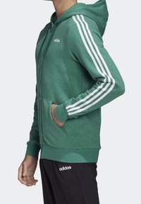 Sudadera verde con cremallera frontal, diseño de tres franjas blancas en las mangas y bolsillos tipo canguro. Fabricada con tejido suave y con capucha ajustable.