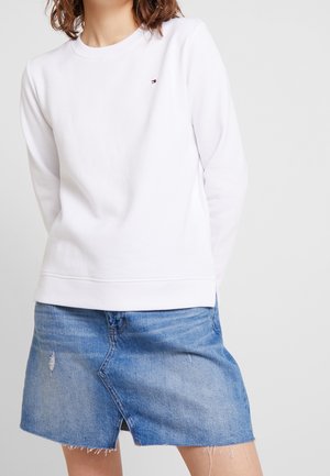 Persona que lleva una sudadera blanca de manga larga lisa combinada con una falda vaquera azul desgastada con una abertura frontal.